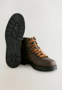 Brun læder vandrestøvle med orange snørebånd, metaløjer, tyk sort sål og dybt mønster, vist fra side- og sålvis.