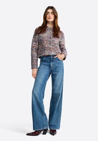 Strukturierte, mehrfarbige kurz geschnittene Strickjacke mit Knöpfen, getragen über hoch taillierten, hellblauen, weit geschnittenen Jeans und spitzen burgunderfarbenen Absätzen.