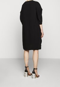 Svart knälång klänning med avslappnad silhuett, med rund halsringning och tre fjärdedels ärmar. Bärs med svarta sandaler och metalliska klackdetaljer.