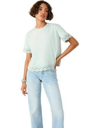 Lyseblå kortærmet t-shirt med blonder vedærmerne og kanten, parret med lyseblå højtaljede jeans. Blød, glat stoftekstur.