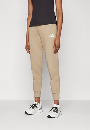 Personne portant un jogging beige Puma, des baskets blanches et noires, et un haut noir sans manches, debout devant un fond clair uni.