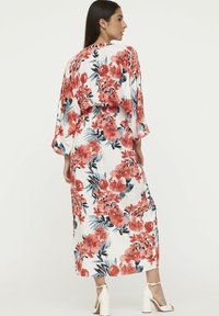Floral Maxi-Kleid mit langen Ärmeln, weißer Stoff mit roten, pinken und blauen Blumen, in der Taille gerafft. Kombiniert mit weißen Blockabsatzsandalen.