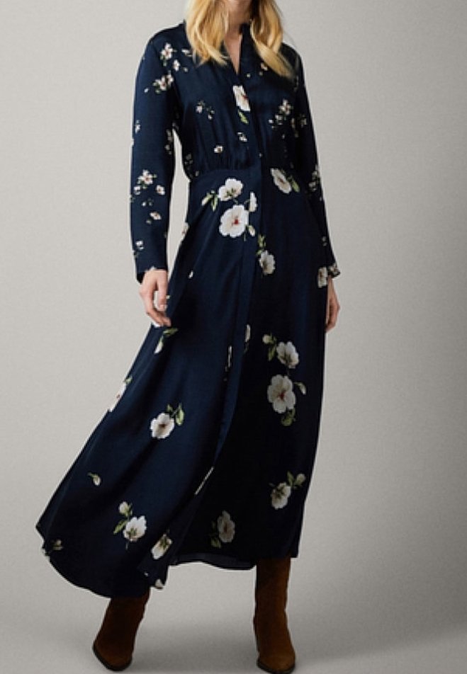 Femme portant une robe maxi à manches longues bleu marine avec des fleurs blanches, associée à des botillons marron, se tenant devant un fond uni.