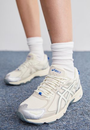 Nogi w białych skarpetkach i beżowych butach sportowych Asics stojące na niebieskim dywanie.