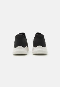 Chaussures de sport noires avec une tige texturée, semelle extérieure blanche et col rembourré. Design minimaliste avec un logo discret sur le côté.