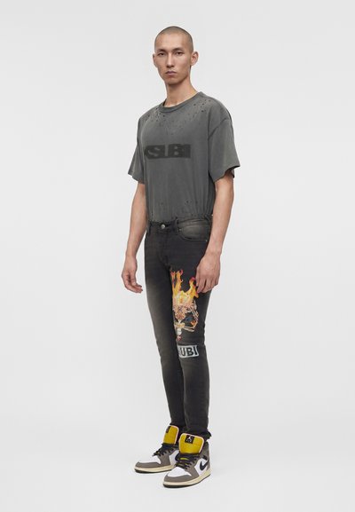 Άνδρας που φοράει μια φθαρμένη σκούρα γκρι μπλούζα με το κείμενο «SIBI», μαύρα τζιν με κέντημα φλεγόμενου κρανίου και μαύρα, λευκά και κίτρινα sneakers πάνω από τον αστράγαλο.