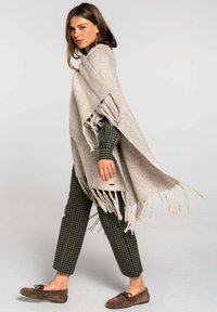 Poncho de punto color beige con bordes con flecos, llevado sobre una camisa a cuadros en verde oscuro y zapatos mocasín marrones. Diseño suelto y texturizado.