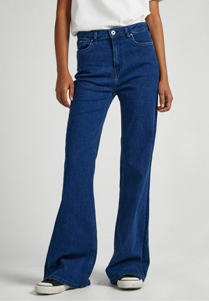 Persoon die donkerblauwe hoge taille flare jeans en zwart-witte sneakers draagt, staand tegen een effen lichte achtergrond.
