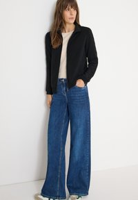 Giacca nera con zip e colletto, abbinata a jeans blu a gamba larga. L'outfit presenta una maglietta di colore neutro sotto.