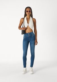 Top halter blanco corto con cierre frontal, jeans skinny azules de cintura alta con detalles de botones, zapatillas blancas y gafas de sol rosas.