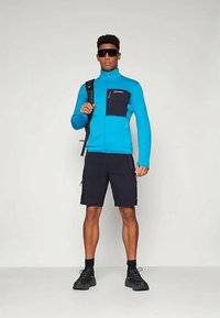 Modra flis jakna s črnim žepom na prsih, v kombinaciji s črnimi kratkimi hlačami. Črna nahrbtnik in sončna očala dopolnjujeta praktično zunanjo obleko.