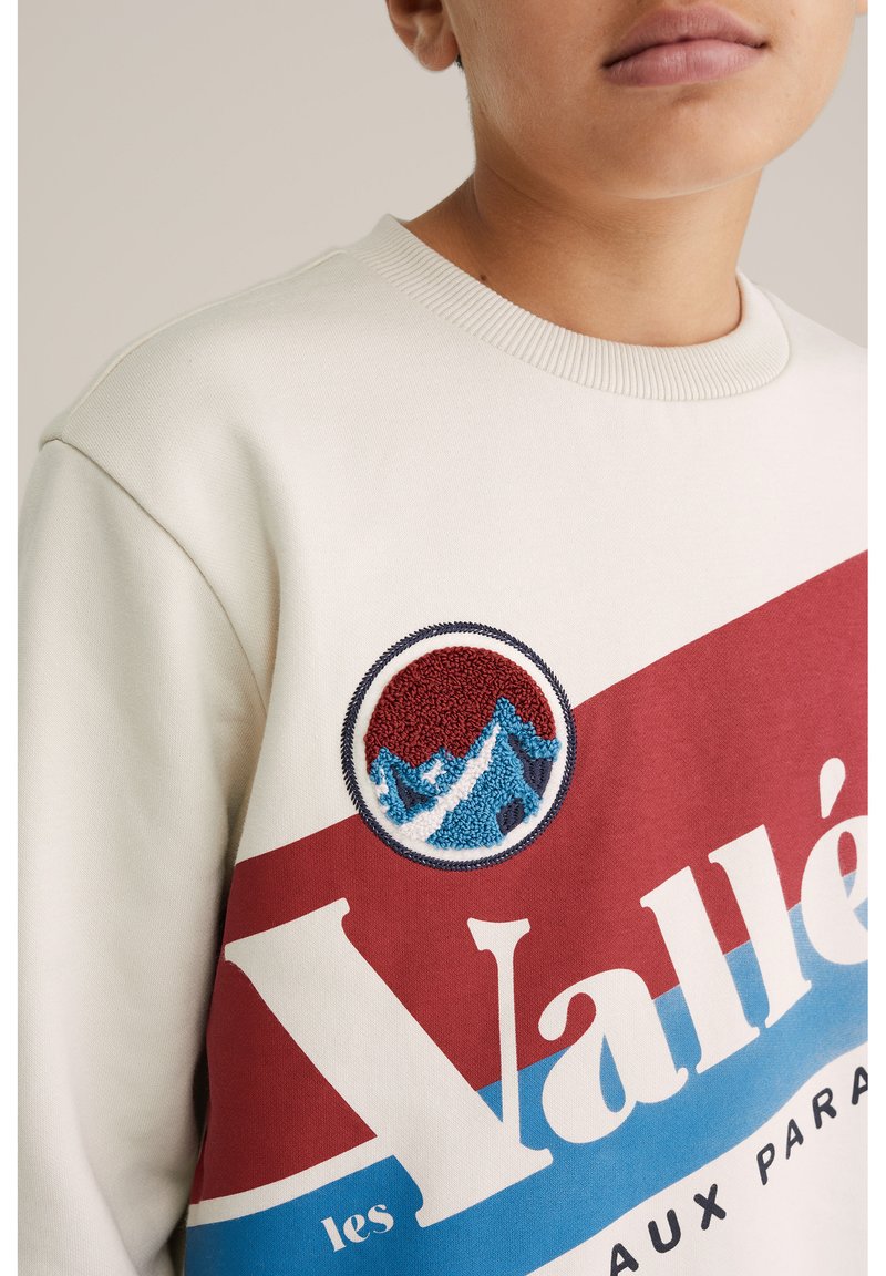 Crew neck sweatshirt i grädde med grafisk design i röd och blått. Har en broderad rund patch med berg och solnedgångsdetalj.