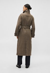 Mulher com cabelo apanhado em coque, vestindo um casaco comprido castanho com cinto e gola alta, calças pretas e sapatos pretos, de pé de costas para um fundo simples.