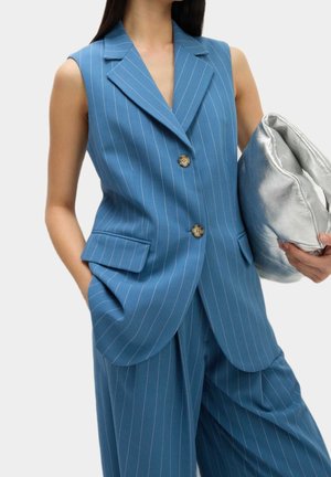 Ermeløs blå blazer med nålestriber og matchende bukser med vide ben, iført og holder en sølvfarvet metallic clutch i den ene hånd.