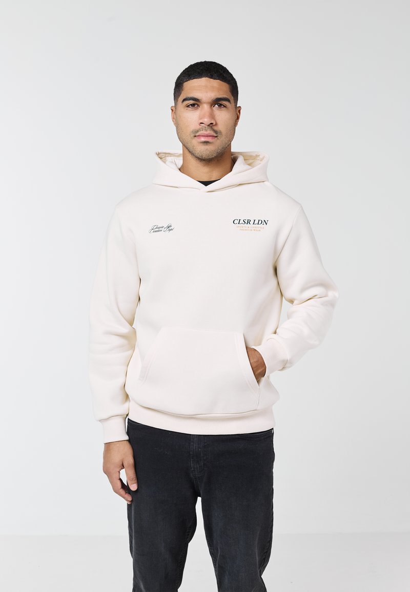 Man som bär en krämfärgad hoodie med framficka och svart text, stående med ena handen i fickan mot en enkel vit bakgrund.