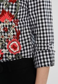 Svart och vit rutigt skjorta med färgglad blommig broderi framtill, med uppvikt ärmar och en knapprad vid manschetten.