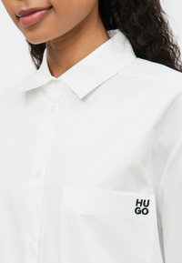 Cămașă albă cu guler clasic, prevăzută cu un buzunar cu "HUGO" brodat în negru. Textură de material neted, design cu nasturi.