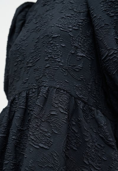 Svart teksturert kjole med preget blomster mønster, med innsvingt midje og flytende silhuett. Myk, rik tekstur.