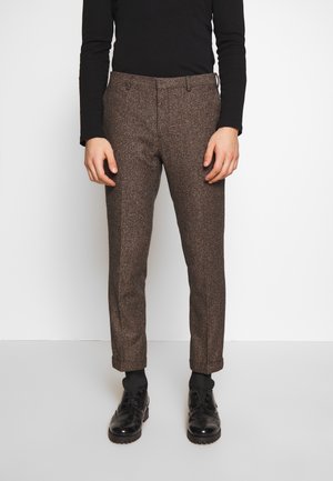 Man wearing brown herringbone patterned trousers, black long-sleeve top, black socks, and black leather dress shoes, standing against a white background.