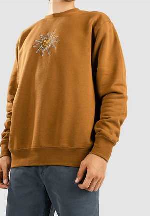 Homme portant un sweat-shirt marron avec un motif de soleil et le texte "HUF" sur la poitrine, associé à un pantalon bleu foncé.