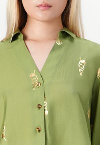 Frau trägt ein grünes Hemd mit Knöpfen und Kragen sowie glänzenden goldenen Muschel-Mustern.