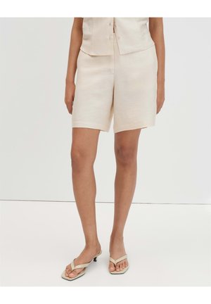 Femme portant un short beige sur mesure et un haut sans manches assorti, associé à des sandales à talons beige clair, debout sur un sol blanc.