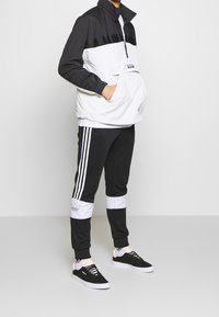 Sporty jacket in black and white, featuring a half-zip, high collar, and front pocket. Paired with black pants with white stripes and sneakers.