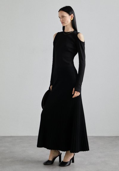 Schwarzes geripptes Strickkleid mit langen Ärmeln und Cutouts an den Schultern, kombiniert mit schwarzen Absatzschuhen und einer runden schwarzen Handtasche.