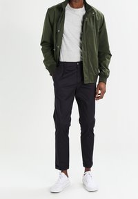 Groene bomberjack, grijs T-shirt, zwarte broek en witte sneakers. Soepele materialen, op maat gemaakte pasvorm en opgerolde broekspijpen.