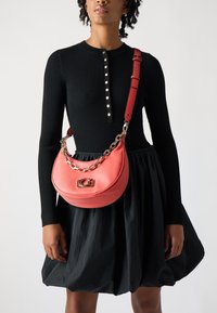 Borsa a spalla in pelle corallo con forma curva, tracolla in catena argentata e chiusura in metallo. Indossata con un top lungo a maniche ribattute nero e una gonna.