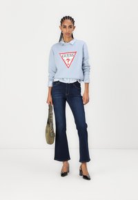 Felpa blu chiaro con logo triangolare rosso, sovrapposta a una camicia a scacchi chiari, abbinata a jeans svasati blu scuro e tacchi neri.