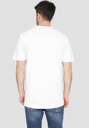 Mann mit kurzem dunklem Haar, der ein schlichtes weißes T-Shirt mit kurzen Ärmeln und blaue Jeans trägt, mit dem Rücken zu einem hellgrauen Hintergrund gewandt.