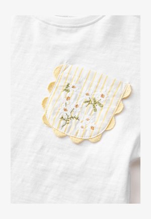 Bela majica s kratkimi rokavi s žepkom z rumenimi črtami in valovitimi robovi, okrašena z vezenimi marjeticami in zelenimi listi.