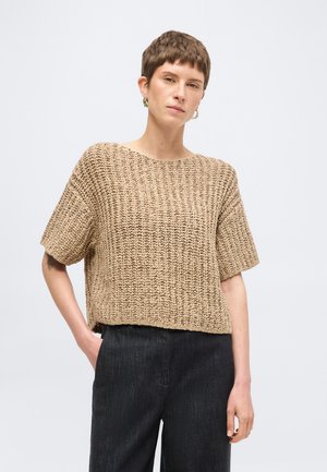 Person with short hair wearing a loose-knit beige short-sleeve sweater and dark high-waisted pants, standing against a plain background.