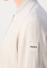 Gros plan d'une personne portant une veste beige clair avec un patch blanc carré portant la mention "BOSS" sur le haut du bras.