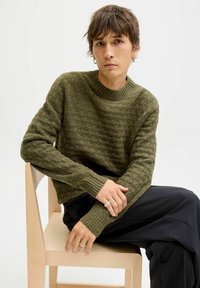 Young person with short brown hair wearing green textured sweater and black pants seated on a light wooden chair against white background.