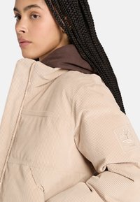 Giacca in corduroy beige con taglio dritto, dotata di un cappuccio marrone sotto. Include un logo ricamato sulla manica sinistra.