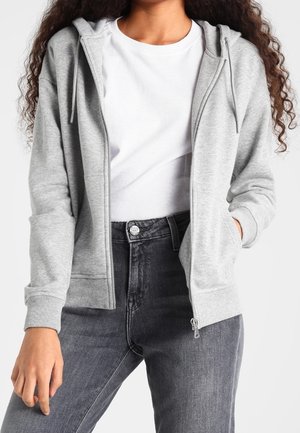 Vrouw draagt een lichtgrijze zip-up hoodie over een wit T-shirt en hoog getailleerde donkergrijze jeans, met haar linkerhand in de zak.