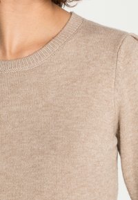 Beige tröja med rund halsringning, strukturerad stickad mönster och lätt ribbing vid kragen. Slät finish med minimalt med detaljer.