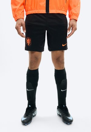 Férfi fekete focishortban, narancssárga oroszlán címkével, fekete Nike zokniban, fekete kék díszítésű stoplis cipőben és narancssárga dzsekiben.