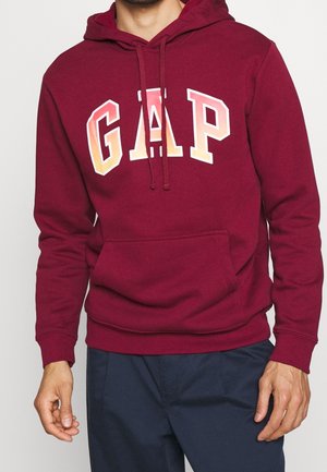 Hombre con sudadera granate con capucha y la palabra "GAP" en letras naranjas y blancas, combinado con pantalones azul marino, de pie con los brazos relajados.