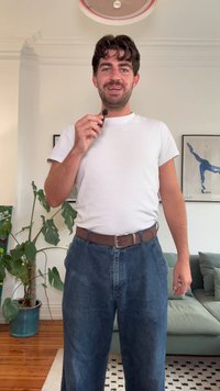 Valkoinen t-paitaa, tummansiniset farkut, ruskea vyö. Sisätila, jossa on vihreä kasvi ja vaaleansävyinen sohva taustalla.