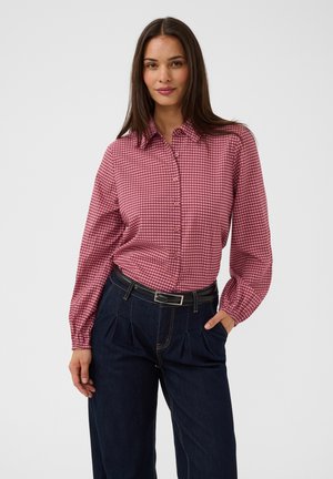 Femme portant une chemise à carreaux rouge et blanche boutonnée, rentrée dans un pantalon taille haute bleu foncé avec une ceinture noire, debout avec une main dans la poche.