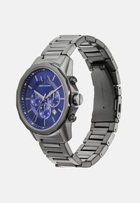 Roostevabast terasest kell, millel on kaheksanurkne korpus, sinine kell Näit, millel on kolm aldialti, hõbedased detailid ja lingiga rihm.