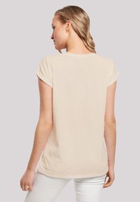 Beige T-Shirt mit kurzen Ärmeln, lockere Passform, weicher Stoff, runder Halsausschnitt, flache Nähte, Rückansicht, getragen mit weißen Hosen.