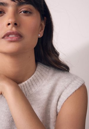 Frau mit mittlerem Hautton, die ein ärmelloses beiges Strickoberteil und kleine Ohrstecker trägt und ihren Arm in der Nähe ihres Gesichts vor einem schlichten Hintergrund anlehnt.