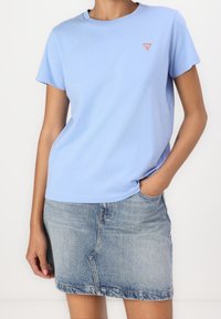 Light blue cotton t-shirt with short sleeves and round neckline, featuring a small red triangular logo on the left chest, paired with a denim skirt.