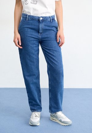 Personne portant un jean bleu taille mi-haute et des baskets blanches, debout sur un sol bleu clair sur fond blanc.