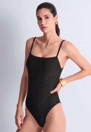Femme portant un maillot de bain une pièce noir texturé avec des fines bretelles, des boucles d’oreilles créoles dorées et un gros bracelet en or, posant avec une main sur la hanche.