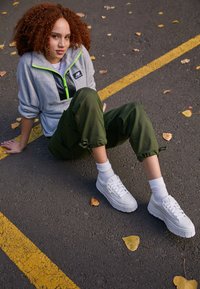 White platform sneakers, gray fleece pullover with black and neon green accents, olive green drawstring pants, sitting on asphalt with leaves.